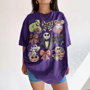 May include: A purple short-sleeve t-shirt featuring a graphic print of characters and bows from The Nightmare Before Christmas. The design includes Jack Skellington, Sally, Zero, Oogie Boogie, and various bows arranged in a grid-like pattern.