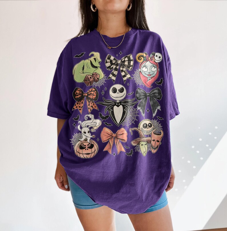 May include: A purple short-sleeve t-shirt featuring a graphic print of characters and bows from The Nightmare Before Christmas. The design includes Jack Skellington, Sally, Zero, Oogie Boogie, and various bows arranged in a grid-like pattern.