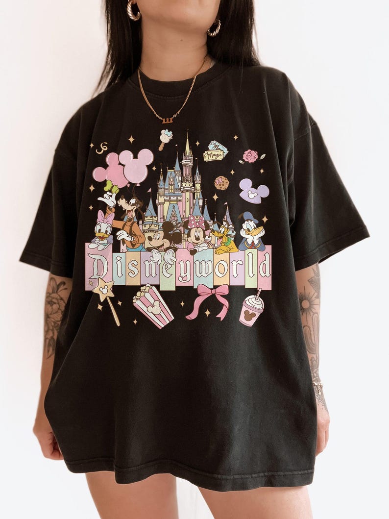 Puede incluir: Camiseta negra con un gr&aacute;fico colorido de Disneyworld. El dise&ntilde;o incluye un castillo, Mickey Mouse, Minnie Mouse, Donald Duck y otros personajes. La palabra "Disneyworld" se muestra en colores pastel.