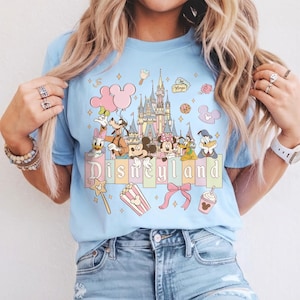 May include: Light blue t-shirt featuring a Disneyland graphic. The design includes Disney characters, a castle, and the word "Disneyland" in pastel colors. Additional elements include balloons, popcorn, and a bow.