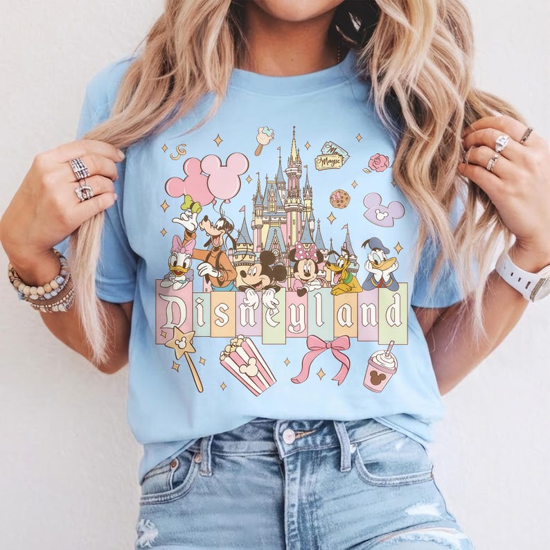 May include: Light blue t-shirt featuring a Disneyland graphic. The design includes Disney characters, a castle, and the word "Disneyland" in pastel colors. Additional elements include balloons, popcorn, and a bow.