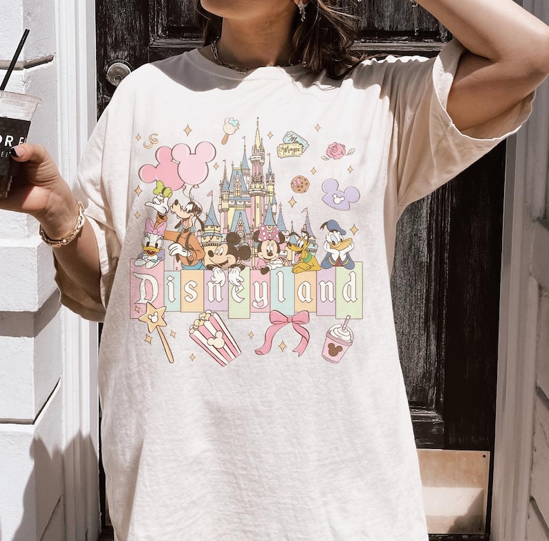 May include: Cream-colored t-shirt featuring a colorful Disneyland graphic. The design includes cartoon characters, a castle, and the word "Disneyland" in pastel colors. Additional elements include popcorn, a bow, and a drink.