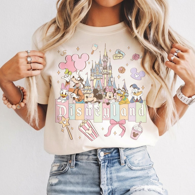 May include: Cream-colored t-shirt featuring a Disneyland graphic. The design includes Mickey and Minnie Mouse, Donald Duck, and other Disney characters, along with a castle and the word "Disneyland" in pastel colors. Additional elements include popcorn, a bow, and a magic wand.
