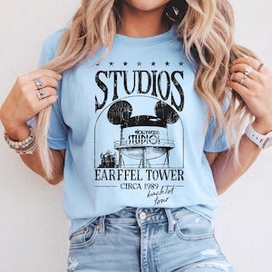 May include: Light blue t-shirt with a black graphic featuring the word "STUDIOS" above a Mickey Mouse-shaped water tower design. Below the tower, the text reads "EARFFEL TOWER CIRCA 1989 backlot tour."