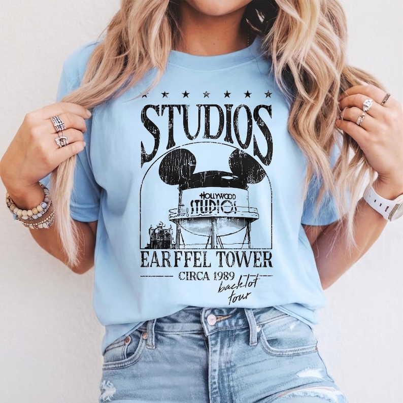 May include: Light blue t-shirt with a black graphic featuring the word "STUDIOS" above a Mickey Mouse-shaped water tower design. Below the tower, the text reads "EARFFEL TOWER CIRCA 1989 backlot tour."