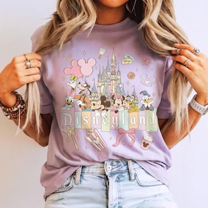 May include: A lilac-colored t-shirt featuring a colorful graphic of Disneyland characters and the Disneyland castle. The design includes Mickey and Minnie Mouse, Donald Duck, and other Disney icons. The word "Disneyland" is printed across the bottom.