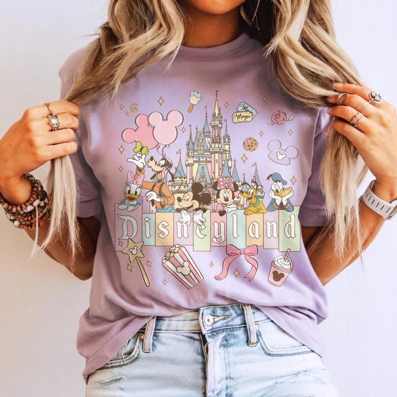 May include: A lilac-colored t-shirt featuring a colorful graphic of Disneyland characters and the Disneyland castle. The design includes Mickey and Minnie Mouse, Donald Duck, and other Disney icons. The word "Disneyland" is printed across the bottom.