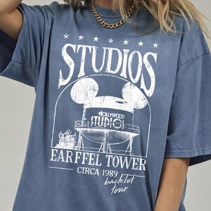 May include: Blue t-shirt with a distressed white graphic featuring the word "STUDIOS" above a Mickey Mouse-shaped water tower design. The text "EARFFEL TOWER CIRCA 1989 backlot tour" is printed below the tower.