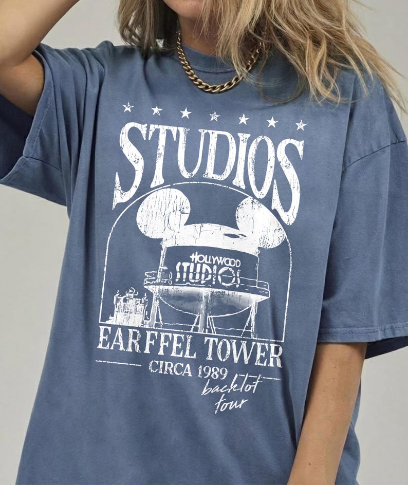 May include: Blue t-shirt with a distressed white graphic featuring the word "STUDIOS" above a Mickey Mouse-shaped water tower design. The text "EARFFEL TOWER CIRCA 1989 backlot tour" is printed below the tower.