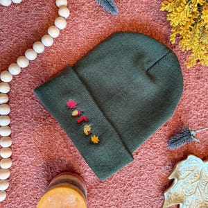Puede incluir: Un gorro verde oscuro con hojas bordadas con temática otoñal y las letras "O, T, T" en rojo, amarillo y naranja. El gorro está doblado en el borde y descansa sobre una superficie texturizada de color naranja rojizo. Elementos decorativos: cuentas de madera y una vela.