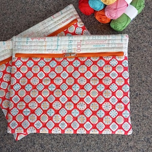 May include: Two quilted fabric pouches with orange zips. The pouches have a red and white geometric pattern on the front and a cream-coloured top with text. Colourful yarn skeins are in the background. The pouches are handmade.