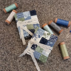 May include: Two patchwork fabric needle cases with lace trim, surrounded by spools of thread in various colours, including blue, green, and grey. Also, there are several pins with blue and white decorative heads. The items are arranged on a textured surface.