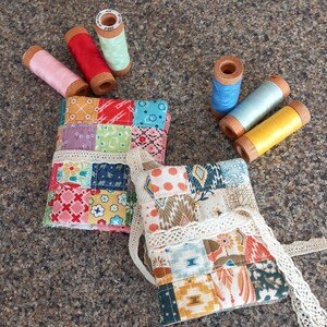 May include: Two patchwork fabric needle cases with lace trim, alongside spools of pink, red, mint green, blue, light blue, and yellow thread. The cases feature a variety of colourful fabric squares, showcasing a handmade, craft theme.
