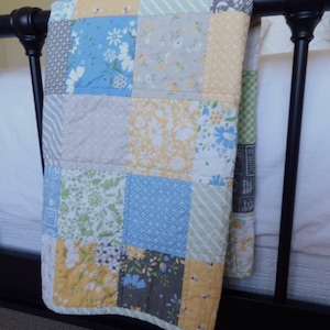 May include: A patchwork quilt draped over a black bed frame. The quilt features a variety of floral and geometric patterns in shades of blue, yellow, green, and gray. The quilt is made of squares of fabric sewn together.