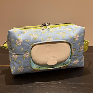 May include: A blue floral patterned wipe dispenser with a light green zipper and trim. The dispenser has a white plastic opening for wipes and a small green handle. The brand name "up&up" is visible on the dispenser.
