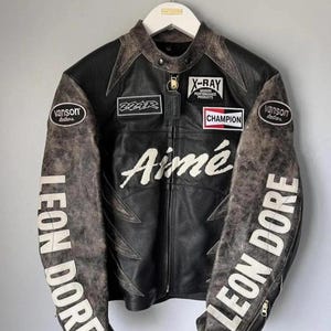 May include: Black leather motorcycle jacket with distressed detailing and patches. Features the text "Aimé" across the chest and "LEON DORE" on the sleeves. Includes patches with "X-RAY", "CHAMPION", and "vanson leathers" branding.