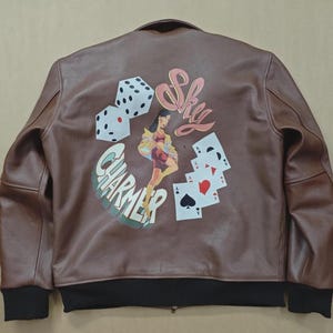 May include: Brown leather bomber jacket with black ribbed cuffs and hem. The back features a graphic with dice, playing cards, a woman, and the words "Sky Charmer".