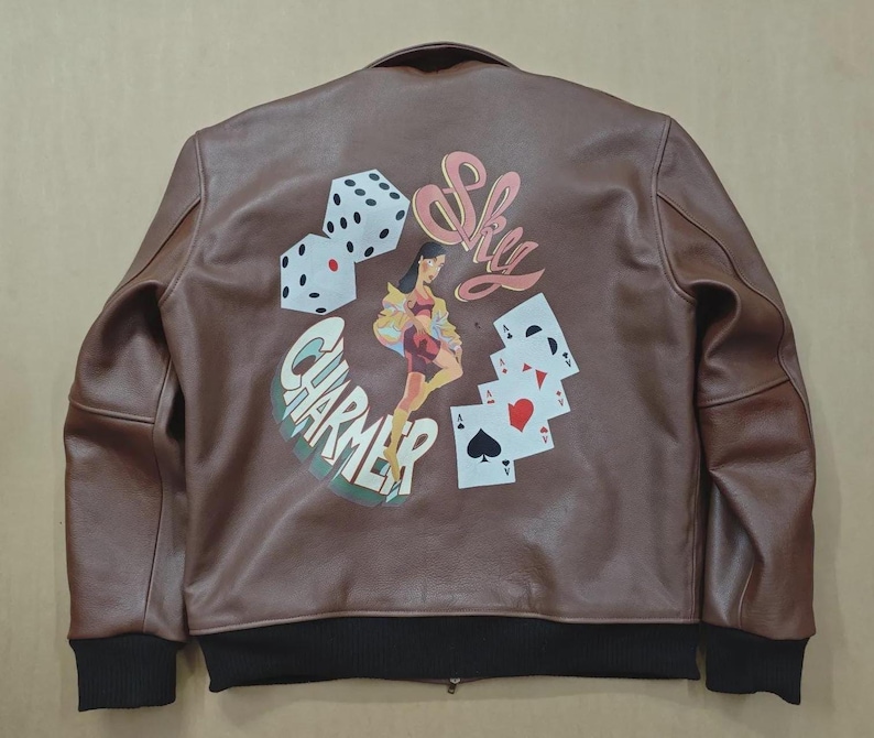 May include: Brown leather bomber jacket with black ribbed cuffs and hem. The back features a graphic with dice, playing cards, a woman, and the words "Sky Charmer".