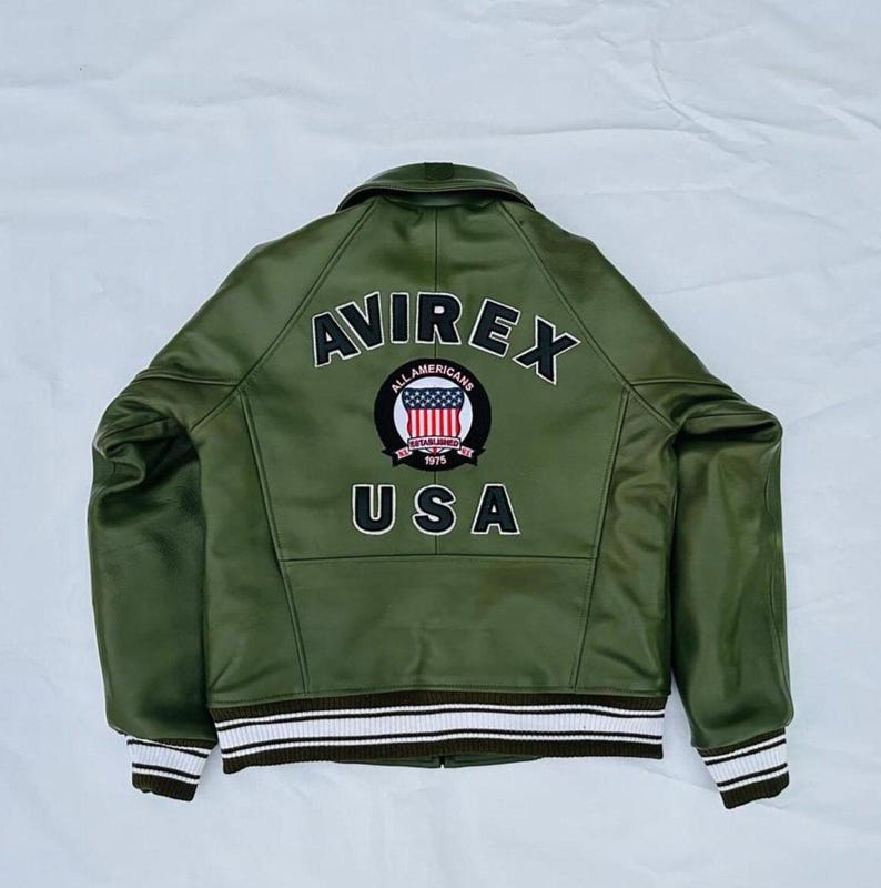 May include: Olive green leather jacket with the word "AVIREX" across the shoulders and "USA" at the bottom. A circular patch in the center features the words "ALL AMERICANS" and a design of the American flag. The jacket has a ribbed collar and cuffs.