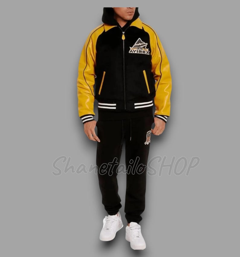 May include: Black and yellow varsity jacket with the "AVIREX" logo on the chest. The jacket has a black hood and is paired with black sweatpants and white sneakers. The outfit is a complete set.