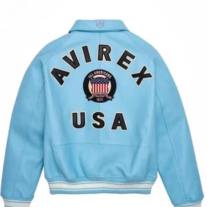 Puede incluir: Chaqueta bomber azul claro con la palabra "AVIREX" en letras negras en la espalda. Debajo, un parche circular con la bandera americana y las palabras "ALL AMERICANS ESTABLISHED 1975". La palabra "USA" est&aacute; debajo.