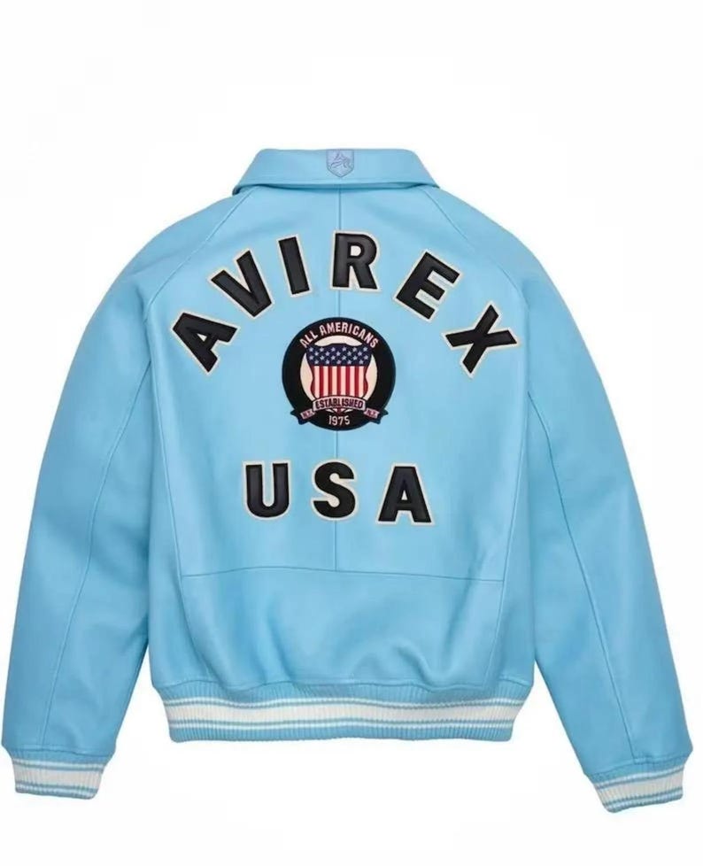 Puede incluir: Chaqueta bomber azul claro con la palabra "AVIREX" en letras negras en la espalda. Debajo, un parche circular con la bandera americana y las palabras "ALL AMERICANS ESTABLISHED 1975". La palabra "USA" est&aacute; debajo.