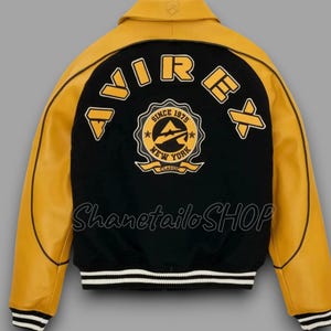 May include: A black and yellow varsity jacket with the word "AVIREX" across the back. The jacket features a patch that says "Since 1975 New York Classic". The jacket has a white and black striped hem and cuffs.