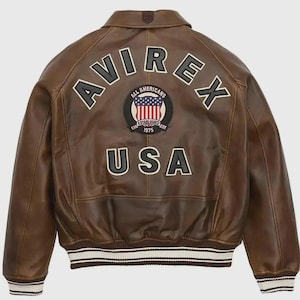 May include: Brown leather jacket with the word "AVIREX" across the shoulders and "USA" at the bottom. A circular patch in the center features the American flag and the words "ALL AMERICANS ESTABLISHED 1975". The jacket has a ribbed white and black striped hem.