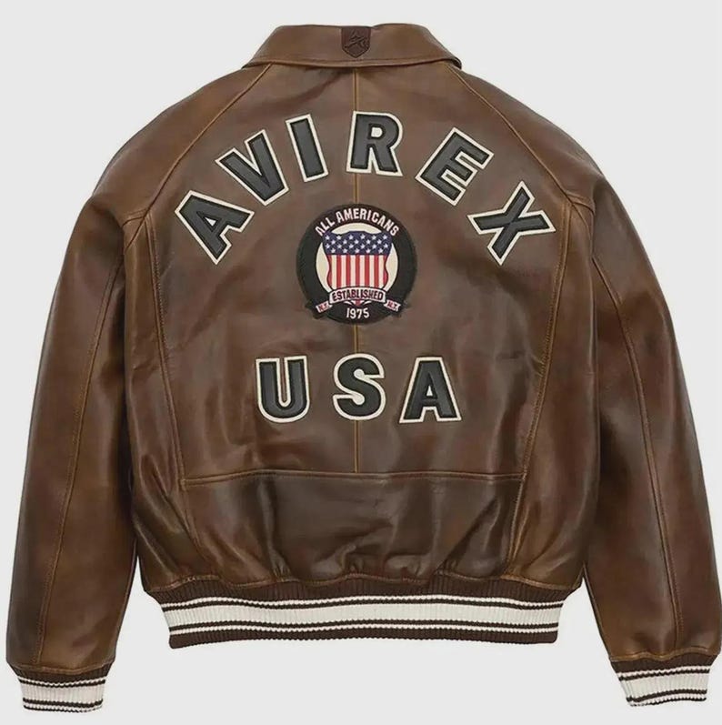 May include: Brown leather jacket with the word "AVIREX" across the shoulders and "USA" at the bottom. A circular patch in the center features the American flag and the words "ALL AMERICANS ESTABLISHED 1975". The jacket has a ribbed white and black striped hem.
