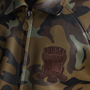 Pu&ograve; includere: Giacca in pelle con motivo mimetico, colletto in pelle marrone e chiusura con cerniera. La giacca presenta una toppa marrone con la scritta "AVIREX" e "ESTABLISHED NY 1975". I colori della giacca includono sfumature di verde, marrone e nero.