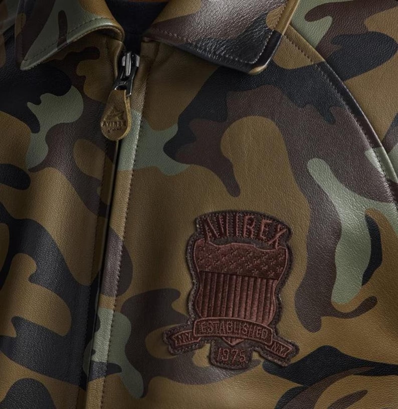 Pu&ograve; includere: Giacca in pelle con motivo mimetico, colletto in pelle marrone e chiusura con cerniera. La giacca presenta una toppa marrone con la scritta "AVIREX" e "ESTABLISHED NY 1975". I colori della giacca includono sfumature di verde, marrone e nero.
