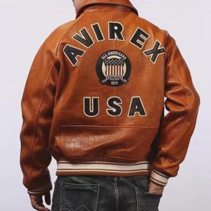Pu&ograve; includere: Giacca in pelle marrone con la scritta "AVIREX" e "USA" sul retro, e un emblema circolare. La giacca ha colletto, polsini e orlo a coste con strisce bianche. La parte posteriore della giacca &egrave; il punto focale.