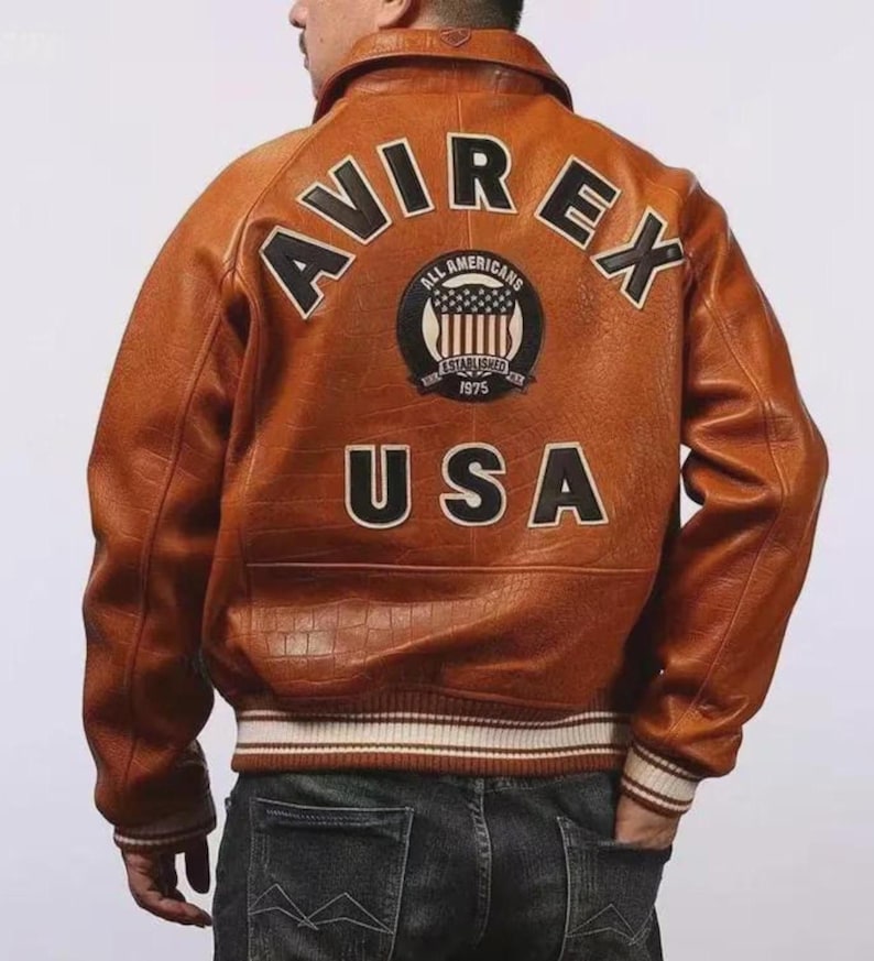 Pu&ograve; includere: Giacca in pelle marrone con la scritta "AVIREX" e "USA" sul retro, e un emblema circolare. La giacca ha colletto, polsini e orlo a coste con strisce bianche. La parte posteriore della giacca &egrave; il punto focale.
