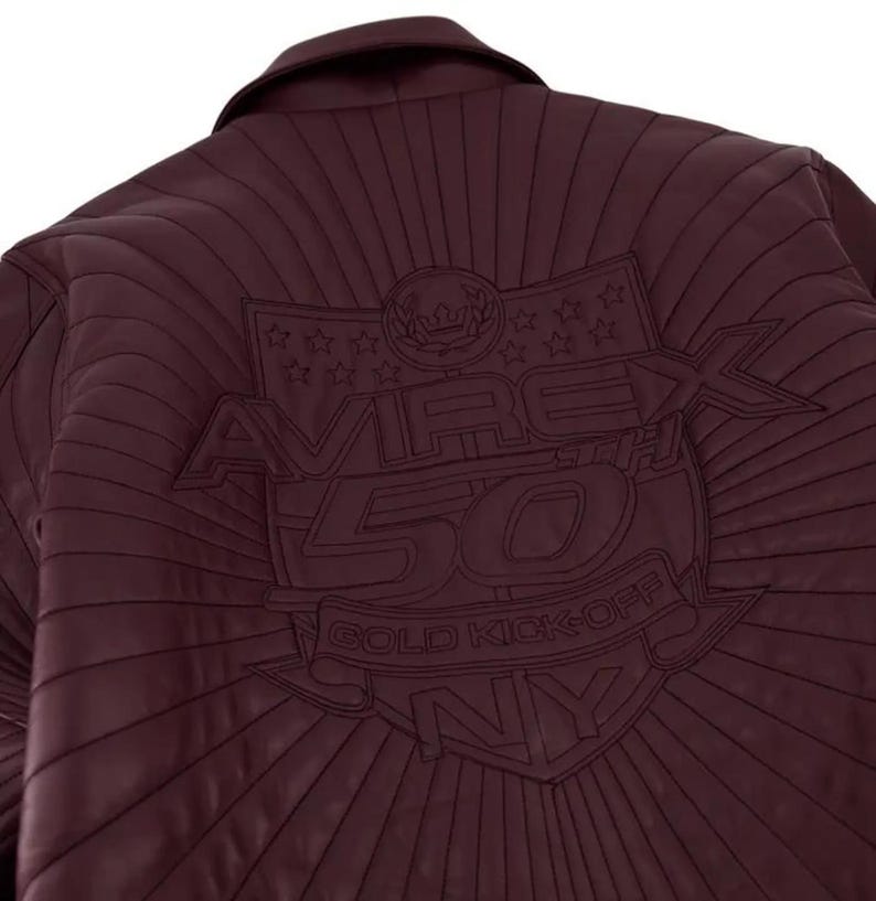 Pu&ograve; includere: Giacca in pelle bordeaux con dettagli cuciti. La schiena presenta un emblema ricamato con la scritta "AVIREX 50TH GOLD KICK-OFF NY" e dettagli a stella e corona. La giacca ha un collo.