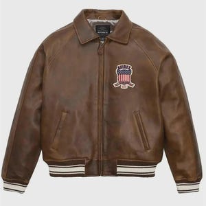 May include: Brown leather jacket with a zip-up front, collared neckline, and a patch on the chest. The jacket features white and black striped cuffs and hem. The patch displays a shield design with stars and stripes.