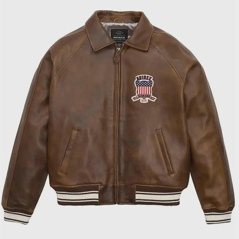 May include: Brown leather jacket with a zip-up front, collared neckline, and a patch on the chest. The jacket features white and black striped cuffs and hem. The patch displays a shield design with stars and stripes.