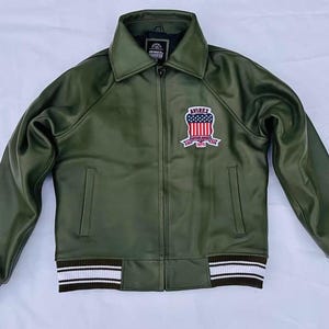May include: Olive green jacket with a collared neckline and a zipper closure. Features a patch on the chest with a red, white, and blue design and the word "AVIREX". The cuffs and hem have white and brown striped detailing.