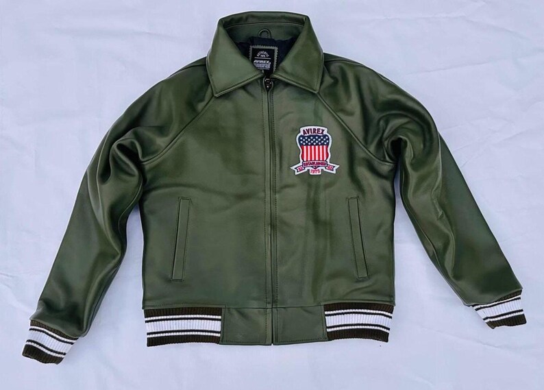 May include: Olive green jacket with a collared neckline and a zipper closure. Features a patch on the chest with a red, white, and blue design and the word "AVIREX". The cuffs and hem have white and brown striped detailing.