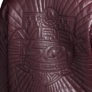 Pu&ograve; includere: Primo piano di una giacca in pelle bordeaux con dettagli cuciti. La parte posteriore presenta un design ricamato con la scritta "AVIREX 50TH GOLD KICK-OFF NY" e motivi a stella e corona d'alloro.