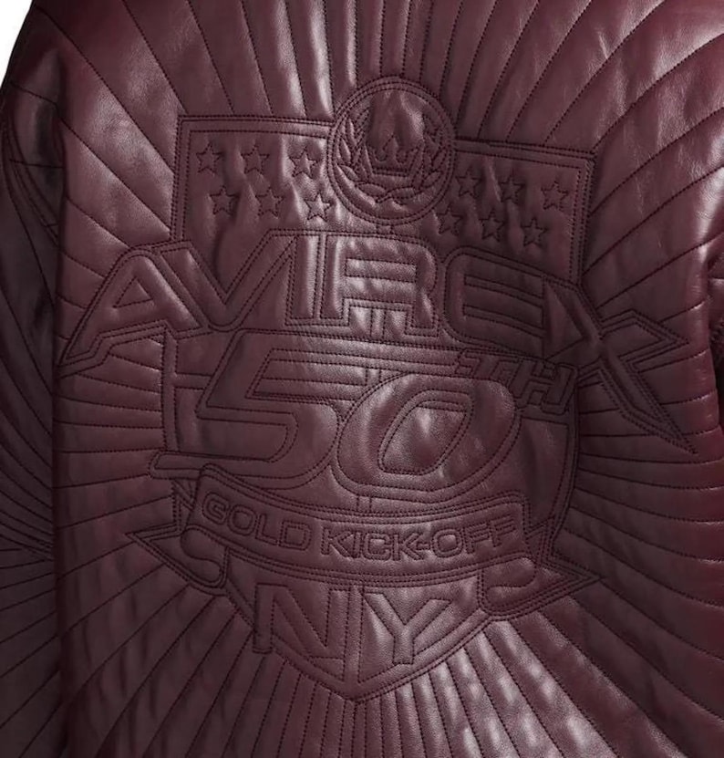 Pu&ograve; includere: Primo piano di una giacca in pelle bordeaux con dettagli cuciti. La parte posteriore presenta un design ricamato con la scritta "AVIREX 50TH GOLD KICK-OFF NY" e motivi a stella e corona d'alloro.