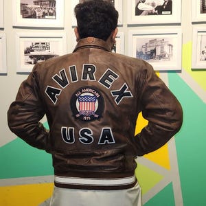 May include: Brown leather jacket with white trim and the text "AVIREX" and "USA" on the back. The jacket features a patch with the words "All Americans" and "Established 1975".