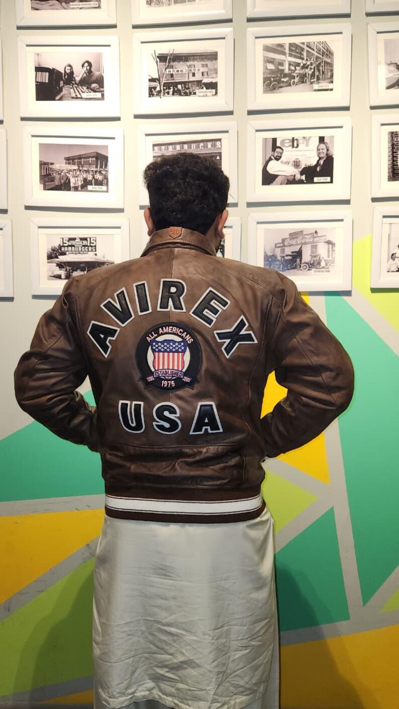 May include: Brown leather jacket with white trim and the text "AVIREX" and "USA" on the back. The jacket features a patch with the words "All Americans" and "Established 1975".