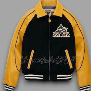 May include: Black and yellow varsity jacket with a zip-up front. The jacket features yellow sleeves and collar, with black on the body. It has a white and black striped hem and cuffs. The jacket has the text "AVIREX" and "1975 Classic" on the chest.