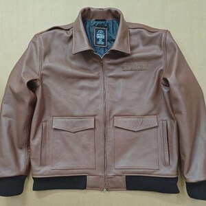 May include: A brown leather jacket with a zip-up front, two front pockets, and a black ribbed hem and cuffs. The jacket has a collar and a small patch on the chest. The lining is dark green.