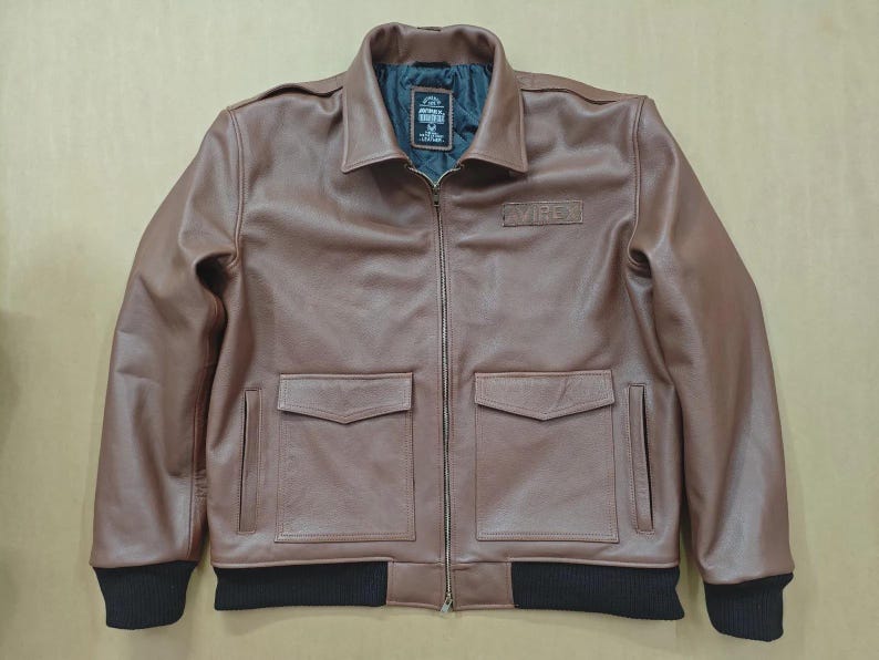 May include: A brown leather jacket with a zip-up front, two front pockets, and a black ribbed hem and cuffs. The jacket has a collar and a small patch on the chest. The lining is dark green.