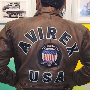 May include: Brown leather jacket with the word "AVIREX" across the back in white lettering. Below the word "AVIREX" is a patch with the words "ALL AMERICANS ESTABLISHED NY 1975 USA". The jacket has a white stripe at the bottom.
