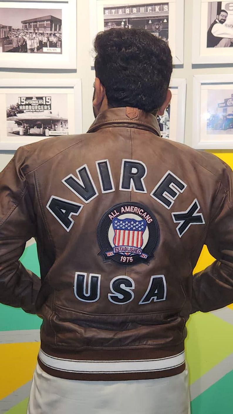 May include: Brown leather jacket with the word "AVIREX" across the back in white lettering. Below the word "AVIREX" is a patch with the words "ALL AMERICANS ESTABLISHED NY 1975 USA". The jacket has a white stripe at the bottom.