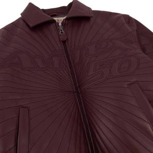 Pu&ograve; includere: Giacca in pelle bordeaux con zip e colletto. La giacca presenta la scritta "AVIREX 50" in un font stilizzato, con linee radiali. La giacca ha un classico stile bomber.