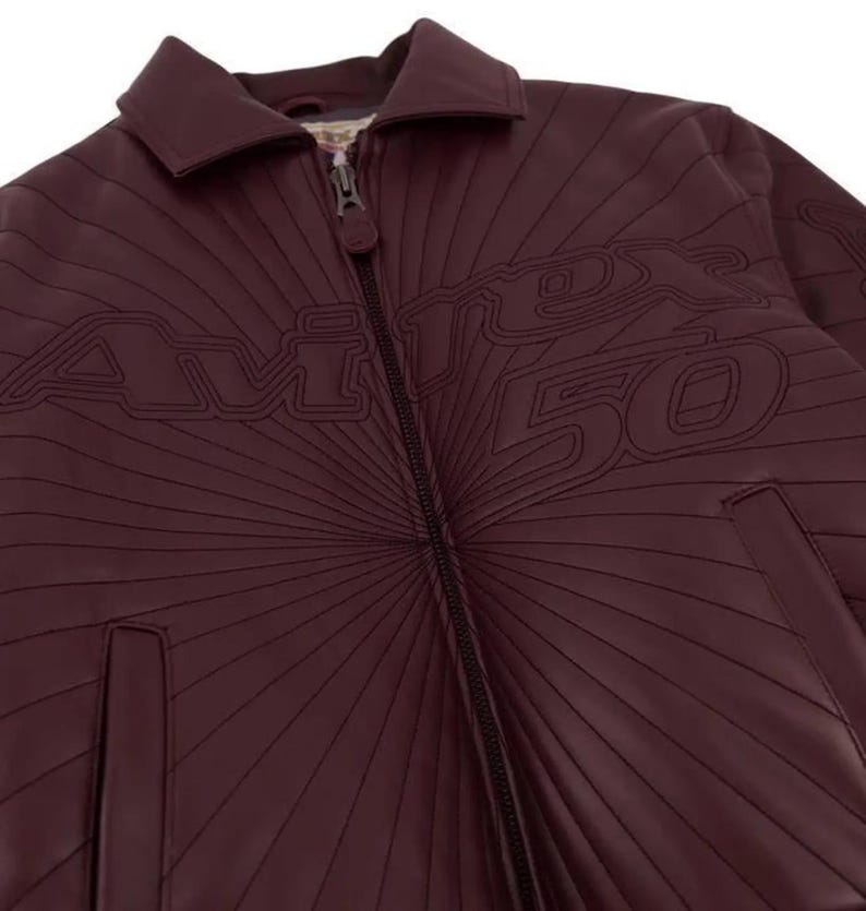 Pu&ograve; includere: Giacca in pelle bordeaux con zip e colletto. La giacca presenta la scritta "AVIREX 50" in un font stilizzato, con linee radiali. La giacca ha un classico stile bomber.