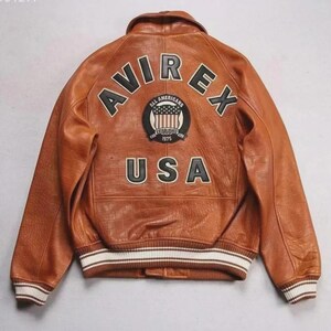 Pu&ograve; includere: Giacca in pelle marrone con la scritta "AVIREX" ad arco sulle spalle e "USA" sulla parte inferiore della schiena. Una toppa circolare con le parole "All Americans" e "Established 1975" &egrave; centrata sopra il testo "USA". La giacca ha polsini a righe bianche e nere.